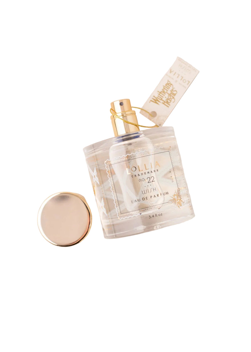 A clear glass Lollia 'WUTHERING HEIGHTS' Wish Eau de Parfum bottle by Margot Elena, with a gold spray nozzle and detached cap, rests on a cream surface next to its blue and gold box—an ode to literary romance.