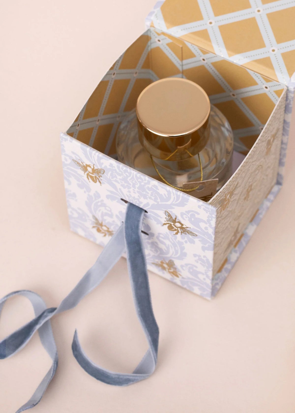 A decorative gold and white box with a blue ribbon holds Margot Elena's Lollia "WUTHERING HEIGHTS" Wish Eau de Parfum, its gold-capped bottle displayed elegantly on a light beige surface.