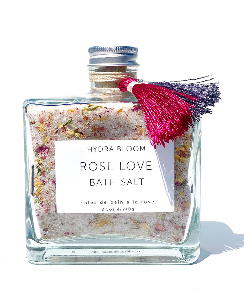 A square glass bottle filled with pink rose-scented bath salts and dried flower petals, labeled "Hydra Bloom Beauty Rose Love Bath Salts," with a decorative red tassel attached to the neck. This
