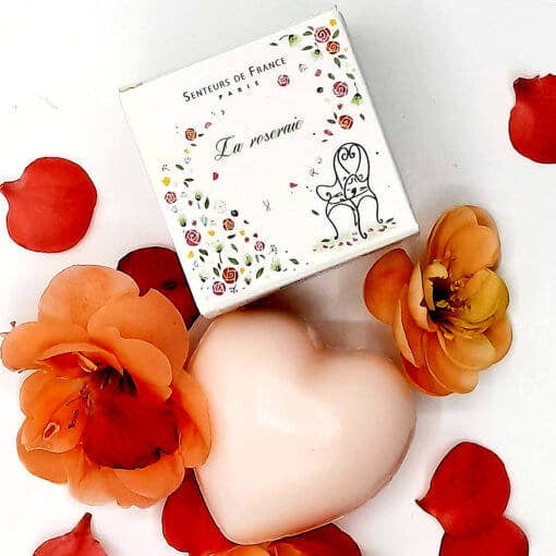 A heart-shaped pink soap surrounded by orange flower petals and a small floral-themed box labeled "Senteurs de France rose garden" on a white background.