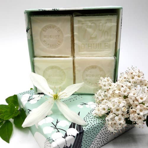 Three bars of Senteurs De France Coffret Jasmine & Muguet Cube Soaps in a decorative box with white and green floral patterns, next to a fresh white flower and baby's breath on a clear surface.