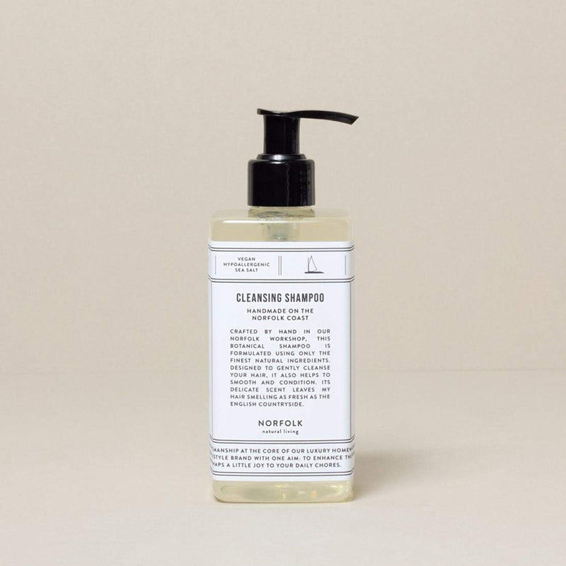 A transparent bottle of Norfolk Natural Living Coastal Cleansing Shampoo for sensitive scalps with a black pump dispenser, against a light beige background. The label details the shampoo's benefits and ingredients.