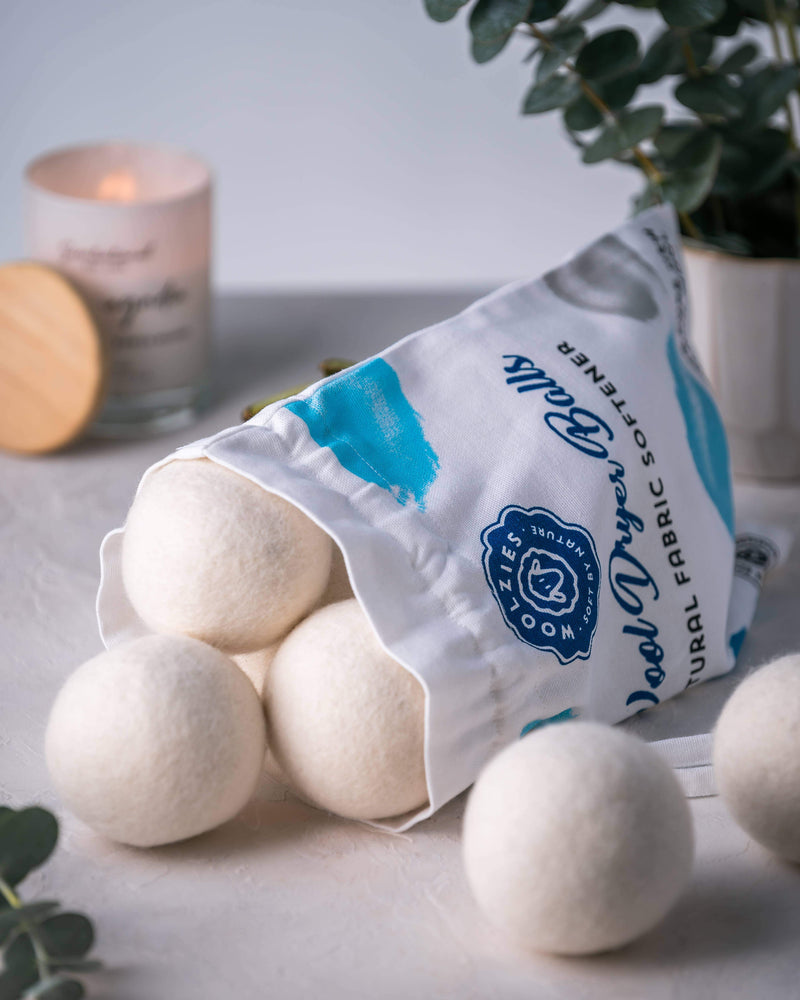 A cloth bag with a logo rests beside the Woolzies Wool Dryer Balls Set of 6 in a Bag made from organic New Zealand wool, with a lit candle and potted plant in the soft-focused background on a light surface.