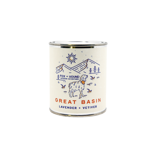 A cylindrical candle in a tin with an illustrated label featuring mountains, trees, a sun, and text "Fox + Hound Lavender + Vetiver National Parks Great Basin Odor Eliminator Soy Candle".