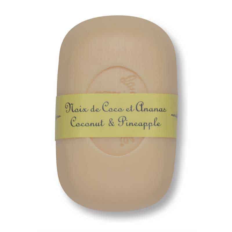 A bar of La Lavande Curved Bar Soap - Coconut & Pineapple - 100gm with a translucent appearance, wrapped in a yellow paper band labeled in French and English "noix de coco et ananas, coconut & pineapple.