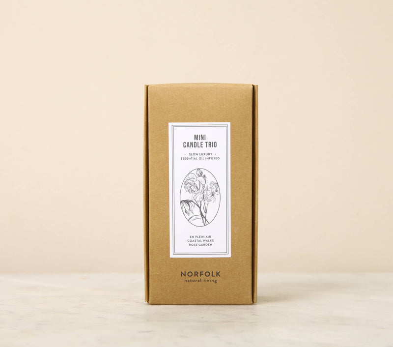 A cardboard box packaging labeled "Norfolk Natural Living - Mini Candle Trio (Coastal, Rose Garden, En Plein Air)" by Norfolk Natural Living, featuring a simple, elegant design with a botanical illustration in the center. The box stands upright against a neutral background.