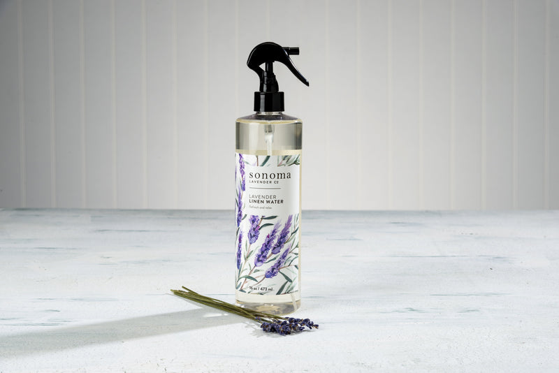 A bottle of Sonoma Lavender Linen Water - 16oz with a spray nozzle, decorated with lavender illustrations. A few lavender stems and buds lie in front on a textured surface, infused with the essential oil of lavender.