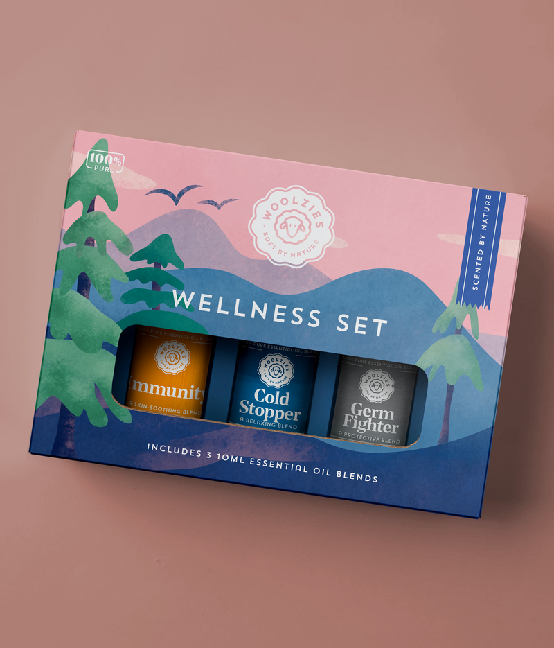 A 'Woolzies The Wellness Essential Blend Oil Collection' box containing three essential oil blends labeled immunity oil blend, cold stopper, and germ fighter, placed against a salmon pink background with green foliage design.