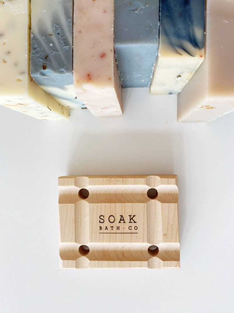 A SOAK Bath Co. soap saver tray with drainage, branded with "SOAK Bath Co," holds a bar of soap, surrounded by various colorful handmade soaps on a white surface.