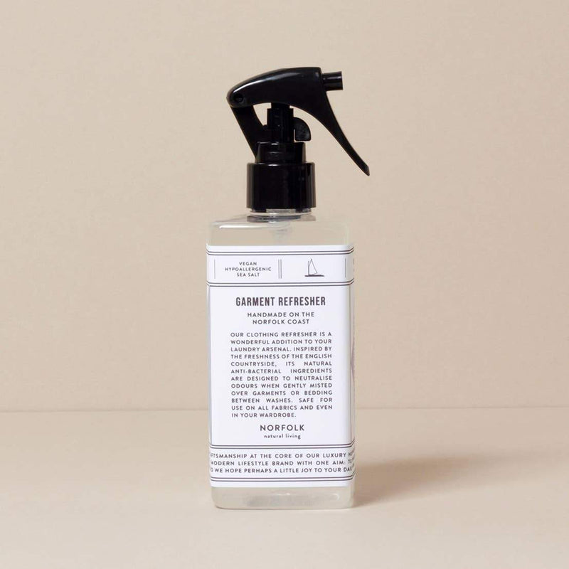 A transparent spray bottle of "Norfolk Natural Living Coastal Garment Refresher" with a white label, detailing its use as a natural fabric refresher with antibacterial properties, against a beige background.