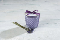 A patterned purple Sonoma Lavender Wax Round Diamond Glass Candle in a glass jar with a silver lid, tied with a purple ribbon and a lavender embellishment, next to dried lavender sprigs on a cracked white surface.