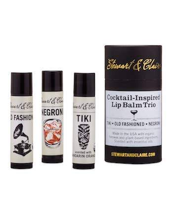 Three Stewart & Claire Cocktail Lip Balm Trio tubes with cocktail-inspired flavors displayed beside their packaging. The packaging highlights flavors like Old Fashioned, Negroni, and Tiki with elegant designs.