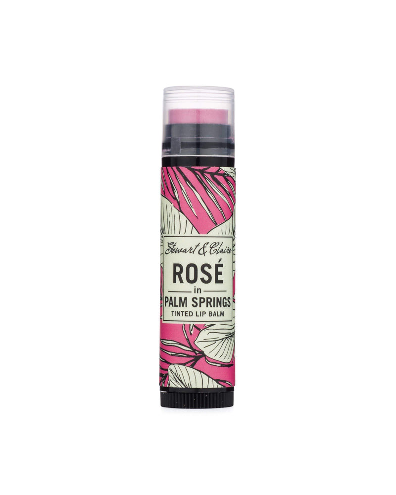 A tube of Stewart & Claire - Rosé In Palm Springs tinted lip balm by stewart & claire featuring a floral and leaf design on the label and crafted with organic ingredients, isolated on a white background