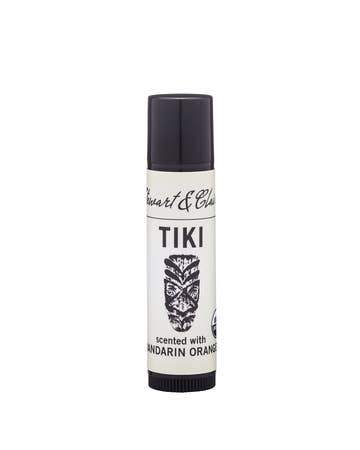 A cylindrical container of Stewart & Claire Tiki Lip Balm with mandarin orange essential oil, featuring a black cap and white label with a tiki mask illustration.