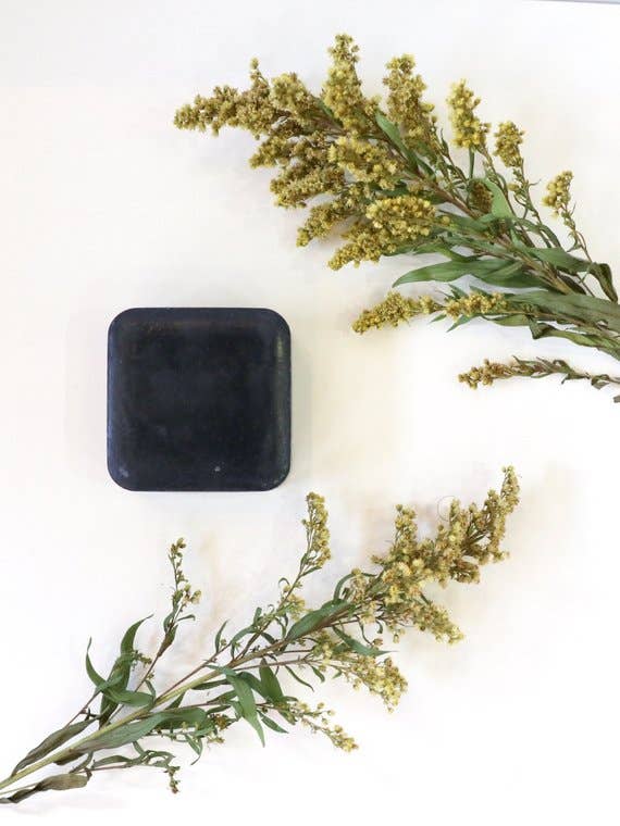 A solid black square container with Homegrown {77833} Co - Cha Cha Charcoal positioned beside sprigs of yellow wildflowers against a plain white background.