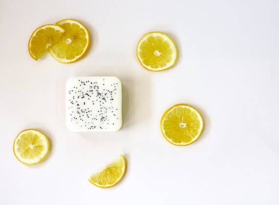 A square bar of Homegrown {77833} Co - My Main Squeeze goat's milk bath bar speckled with black particles, surrounded by fresh orange and lemon slices on a white background.
