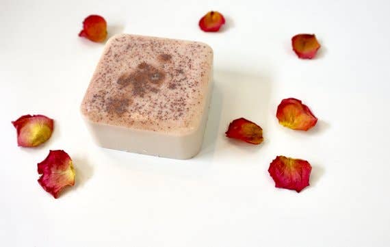 A handmade goat's milk soap bar with pinkish-brown speckles on a white background, surrounded by scattered red rose petals, Homegrown {77833} Co - Rose Colored Glasses.