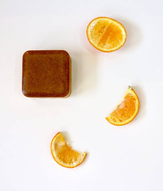 A square brown goat's milk bath bar with three orange slices, one halved and two segments, arranged on a white background from Homegrown {77833} Co - You're the Zest.