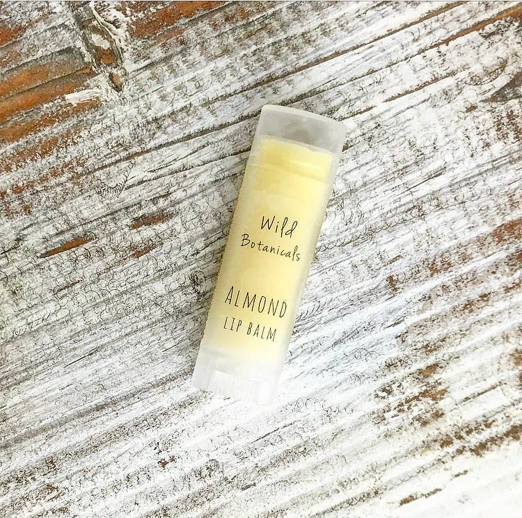 A tube of Wild Botanicals Almond Lip Balm with organic shea butter is placed on a rustic wooden surface. The packaging is simple with clear labeling.