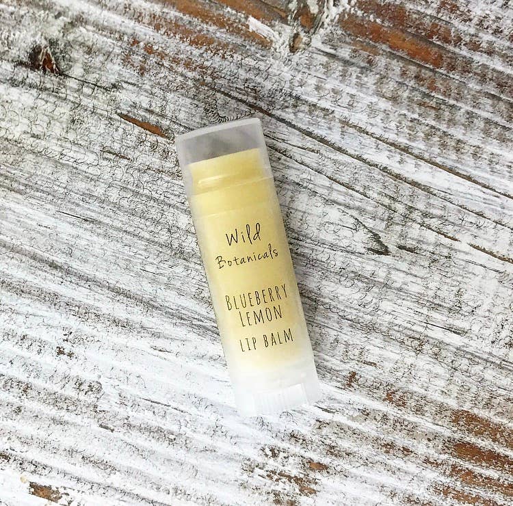 A tube of Wild Botanicals Blueberry Lemon Lip Balm infused with organic jojoba oil, resting on a weathered wooden surface with white paint residue.