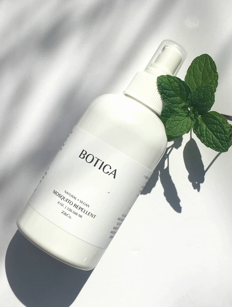 A white bottle of Z&Co. Mosquito Repellent Botica Collection DEET-free spray with mint leaves on its side, casting a shadow on a white surface in bright sunlight.