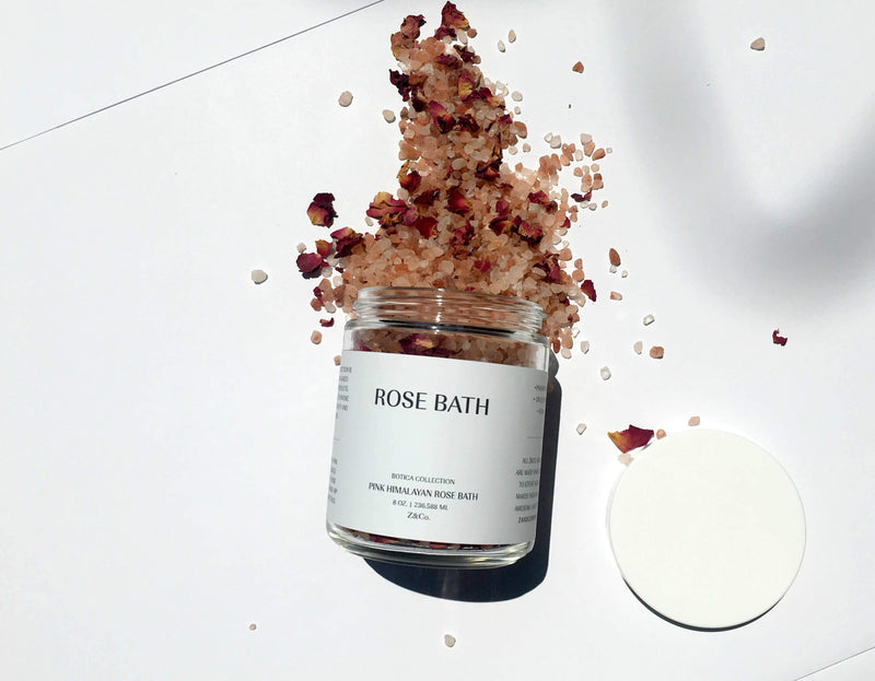 A jar labeled "Z&Co. Pink Himalayan Rose Bath" with pink Himalayan and Dead Sea salts spilling out, mixed with rose petals and a hint of rose geranium essential oil, against a white background.