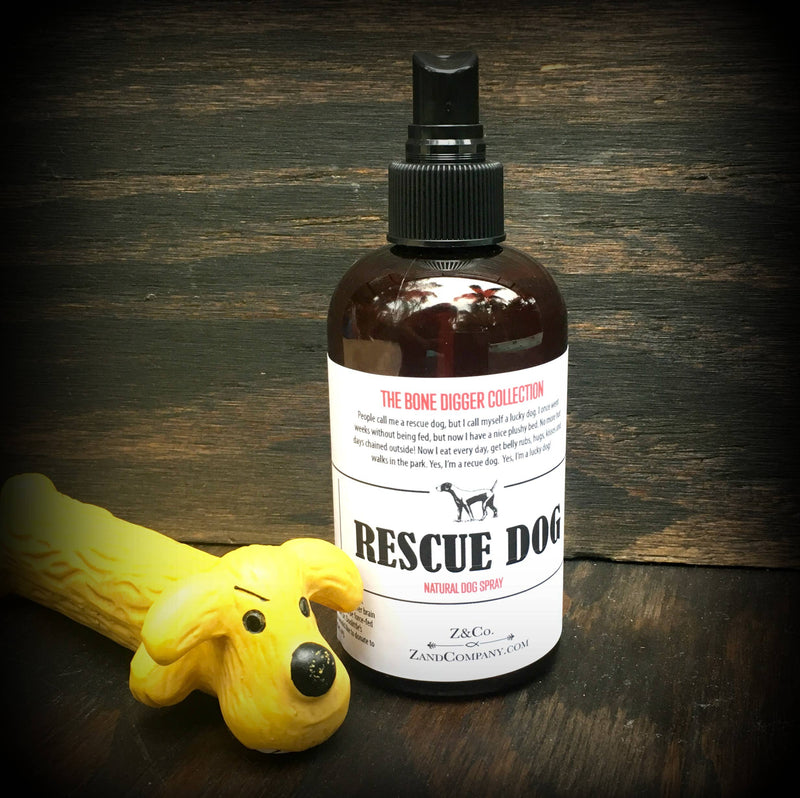 A bottle of Z&Co. Rescue Dog Spray Mist, from the Bone Digger collection, placed on a wooden surface beside a yellow dog-shaped toy. The label features a black and white dog illustration.