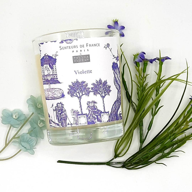 A decorative candle labeled "Senteurs De France Candle Toiles de Jouy Violette" with blue floral designs, accompanied by fresh violet flowers and green leaves on a white background.