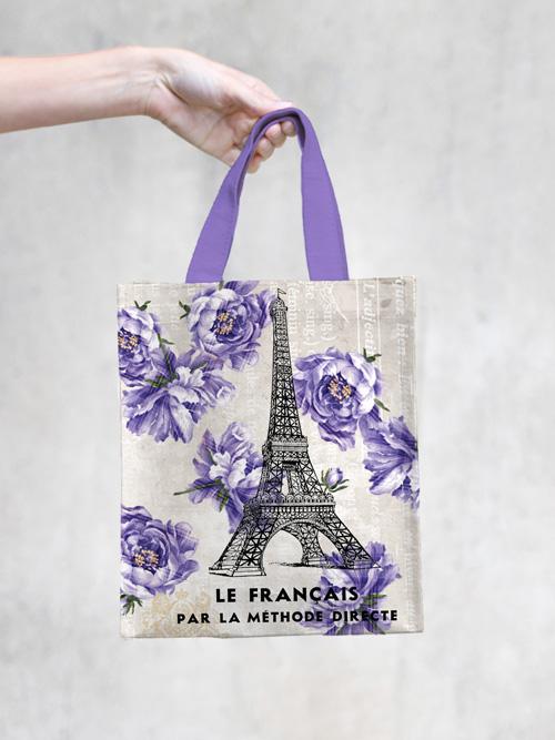 A Margot Elena TokyoMilk Tote Bag - French Kiss Small Tote featuring a design of the Eiffel Tower and purple flowers with the text "le français par la méthode directe," hanging against a plain, lightly textured background. Ideal as a small