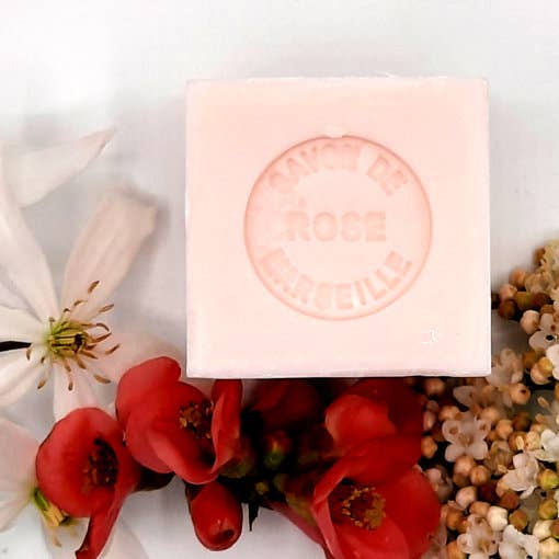 A pink square Senteurs De France Marseille Rose Cube Soap surrounded by red and white flowers and tiny beige blooms on a white background.