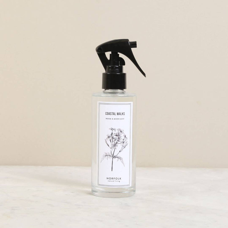 A clear spray bottle labeled "Norfolk Natural Living Coastal Walks Room and Mood Mist - a rosy & fresh blend - norfolk" with an intricate floral sketch, placed on a marble surface against a neutral backdrop.