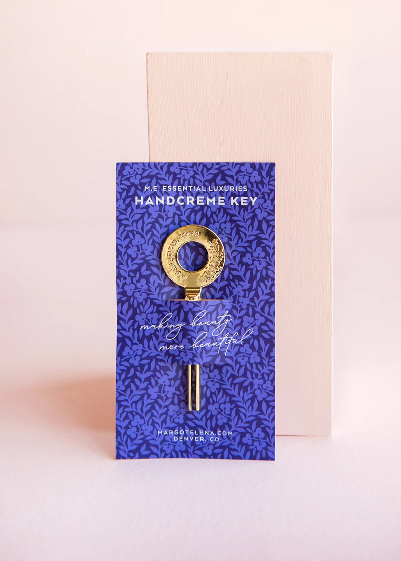 A Margot Elena Essential Luxuries Handcreme Key tube with a luxurious decorative key attached, set against a pink backdrop, emphasizing its elegant packaging design featuring a blue floral pattern.