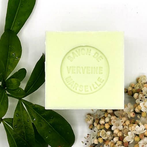 A Senteurs De France Marseille Verbena Cube Soap labeled "savon de lemon verbena" rests on a white surface, surrounded by small dried flowers and fresh green leaves.