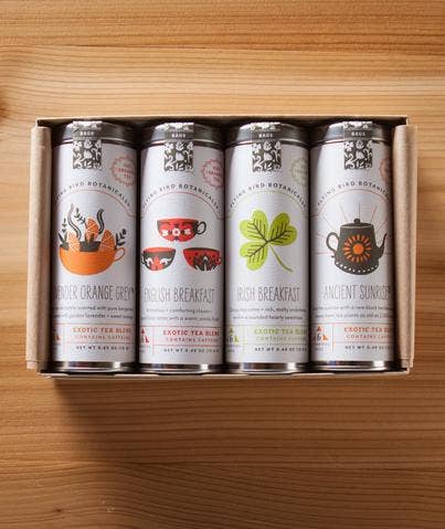 A Flying Bird Botanicals Good Morning Gift Box containing five cylindrical tins of organic loose-leaf tea, each labeled with different flavors including English Breakfast and Irish Breakfast, and featuring colorful, unique designs on a wooden surface.