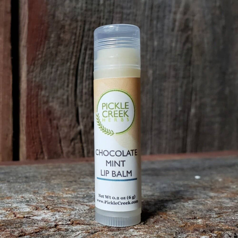 A tube of Pickle Creek Herbs Chocolate Mint Lip Balm displayed against a rustic wooden background. The label is beige with green and brown text.