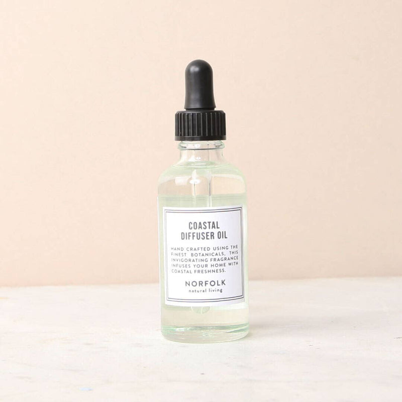 A clear glass bottle of Norfolk Natural Living Coastal Gemstone Oil with a black dropper, labeled with details of handcrafted origins and coastal rose scent, against a light beige background.