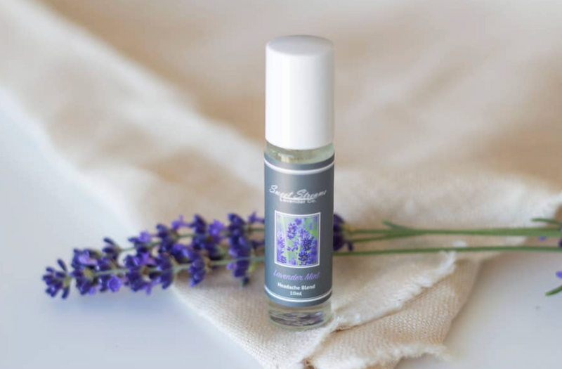 A Sweet Streams Lavender Co. - Lavender Mint Headache Blend Roller sits on a beige cloth next to a sprig of lavender flowers on a white surface. The label features an image of lavender.