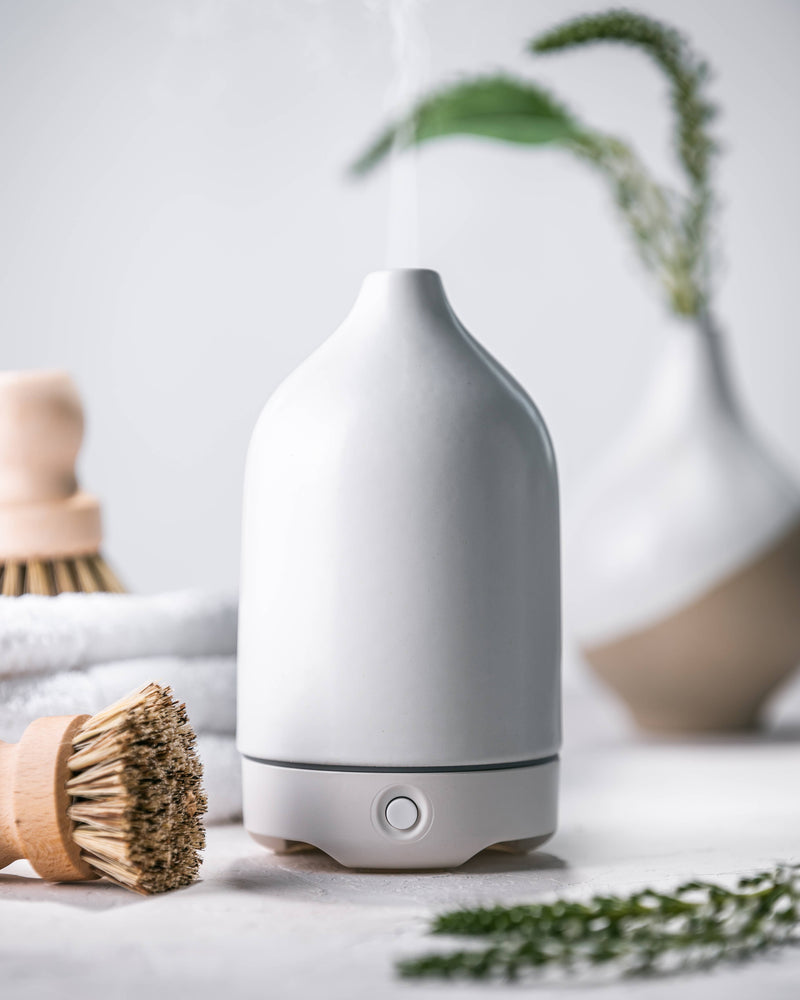 A modern Woolzies White Ceramic Glass Diffuser using ultrasonic technology to release mist, surrounded by spa accessories and green plants, set against a soft, light gray background.