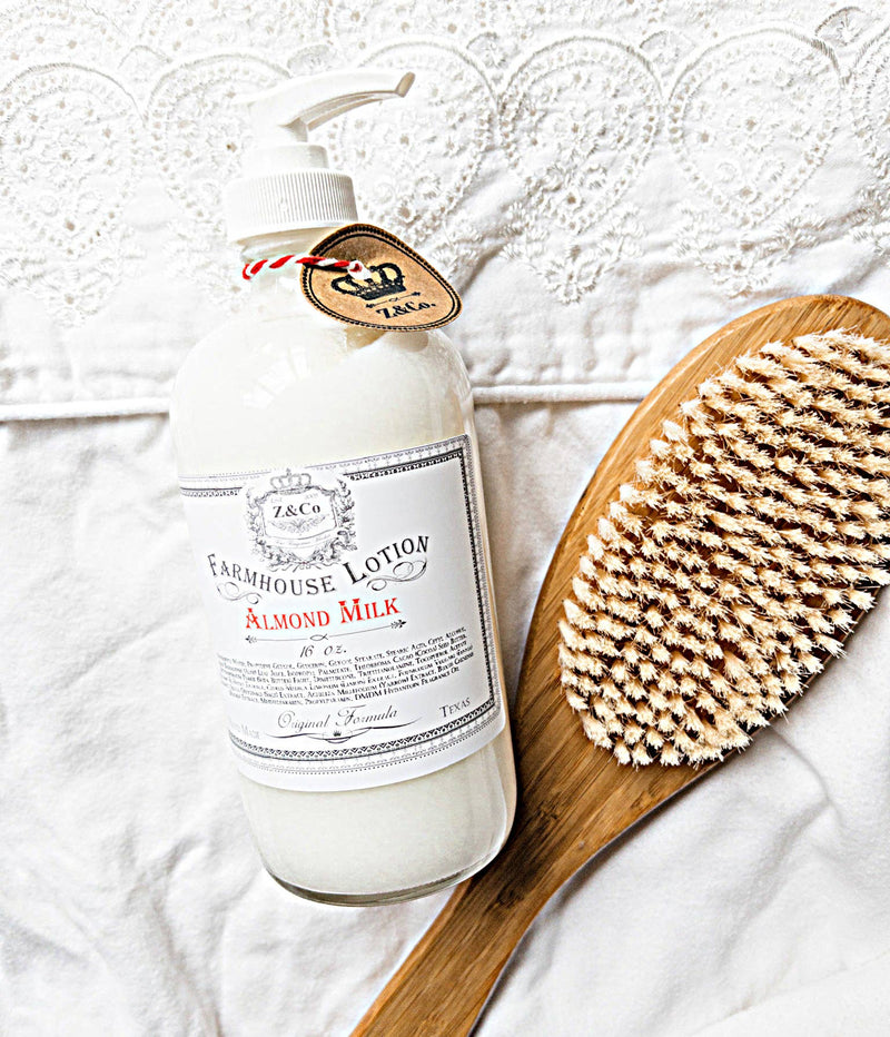 A lotion bottle labeled "Z&Co. Almond Milk Farmhouse Lotion" next to a wooden brush, both on a lace-trimmed white fabric surface.