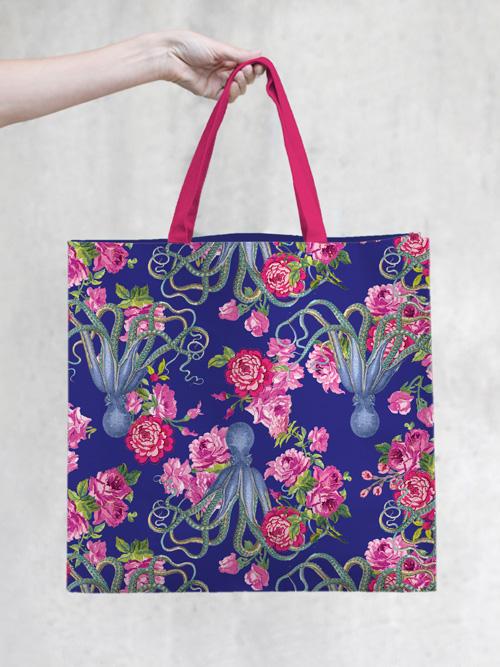 A hand holding a Margot Elena TokyoMilk Tote Bag - 20,000 Flowers Under the Sea Market Tote with a vivid blue background adorned with a floral and octopus print in shades of pink and purple. The bag's woven handles are bright pink.