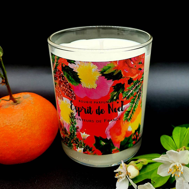 Senteurs De France Candle Cinnamon Spice Scented made of vegetable wax in a glass jar with a vibrant floral label, next to orange and white flowers on a dark backdrop. Text on the candle reads "Esprit de Noël