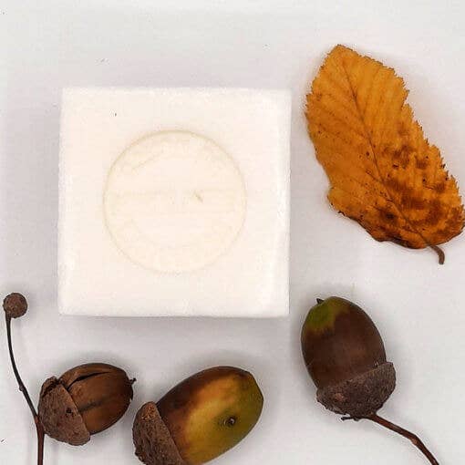A composition featuring natural autumn elements: a leaf, acorns, and a small white cube of Senteurs de France Marseille Bitter Almond Soap with a leaf imprint on a neutral background.