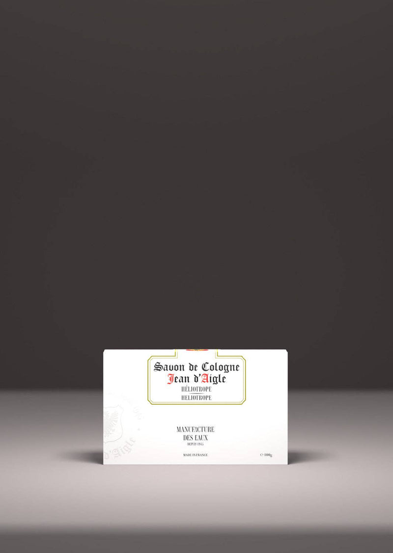 A vintage-style bar of Jean d'Aigle Heliotrope Soap packaging presented on a white pedestal against a gray background. The label features elegant, vintage typography and a classic design.