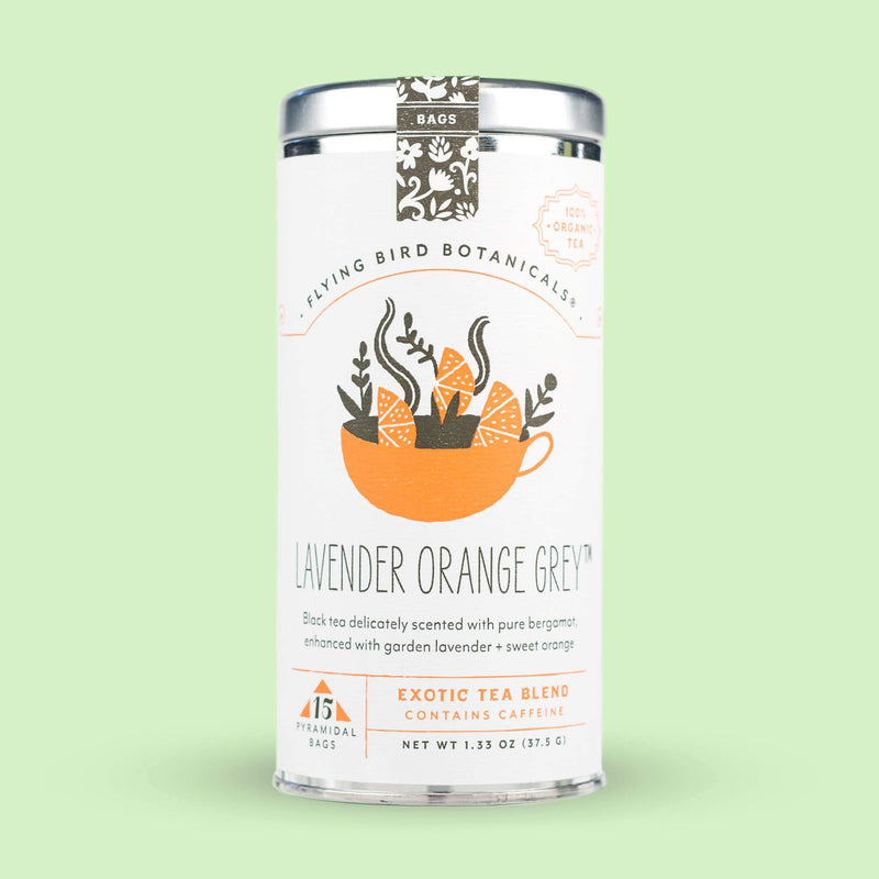 A tin of Flying Bird Botanicals Lavender Orange Grey – 15 Tea Bag Tin against a light green background. The label shows an orange cup with a floral design containing steaming tea.