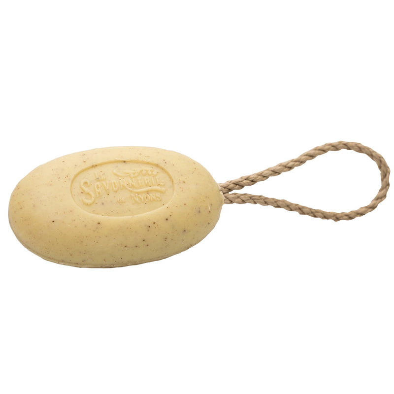 A bar of oval-shaped, light beige La Savonnerie de Nyons Exfoliating Verbena organic shea butter soap with a rustic rope embedded through it for hanging, stamped with the words "savon de marseille." The background is plain.