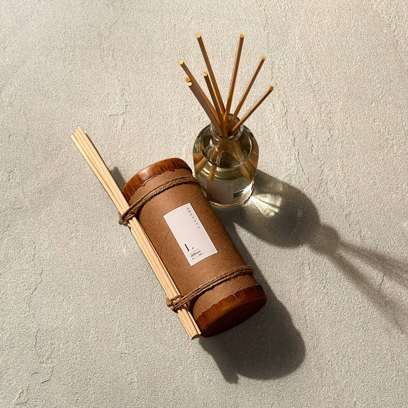 A Lavande fragrance diffuser in a glass container with several bamboo reeds protruding, wrapped in a light brown leather sleeve with a label, placed on a textured surface in sunlight, casting a shadow.