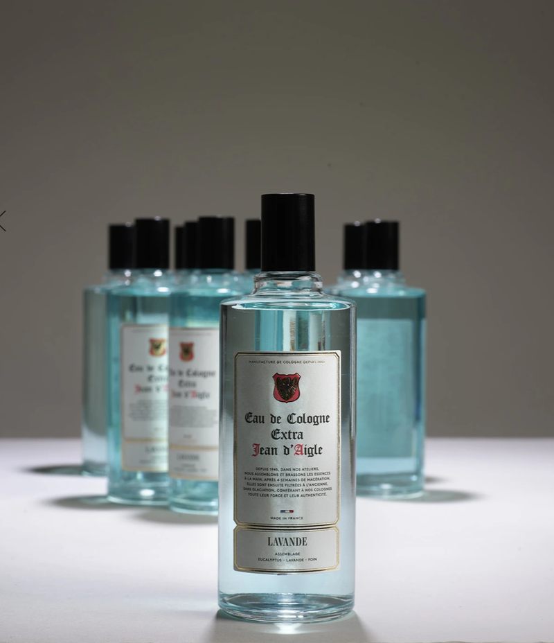 Five clear bottles of Jean d'Aigle Lavender Eau de Cologne, labeled in French, with black caps, are arranged in a spotlight with a soft grey background. The front bottle is in focus while the others are blurry.