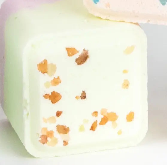 A Lizush Grapefruit Shower Steamer embedded with small orange and yellow dried flower petals, made from vegan ingredients, displayed against a soft pink and white background.