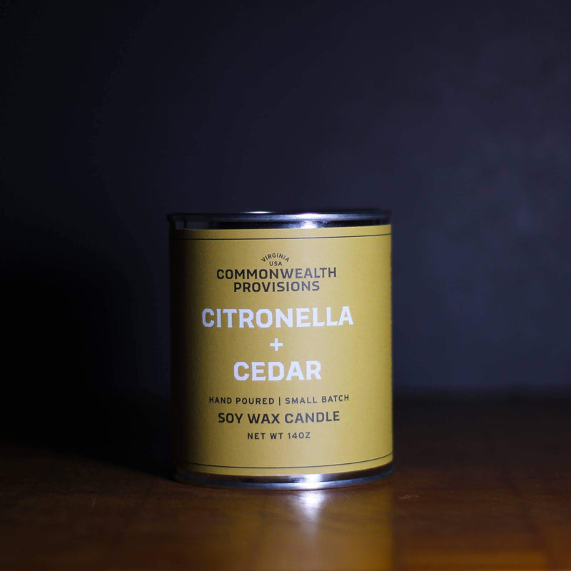 A golden yellow Commonwealth Provisions Citronella + Cedar Candle in a small tin labeled "commonwealth provisions" on a dark background with soft lighting.