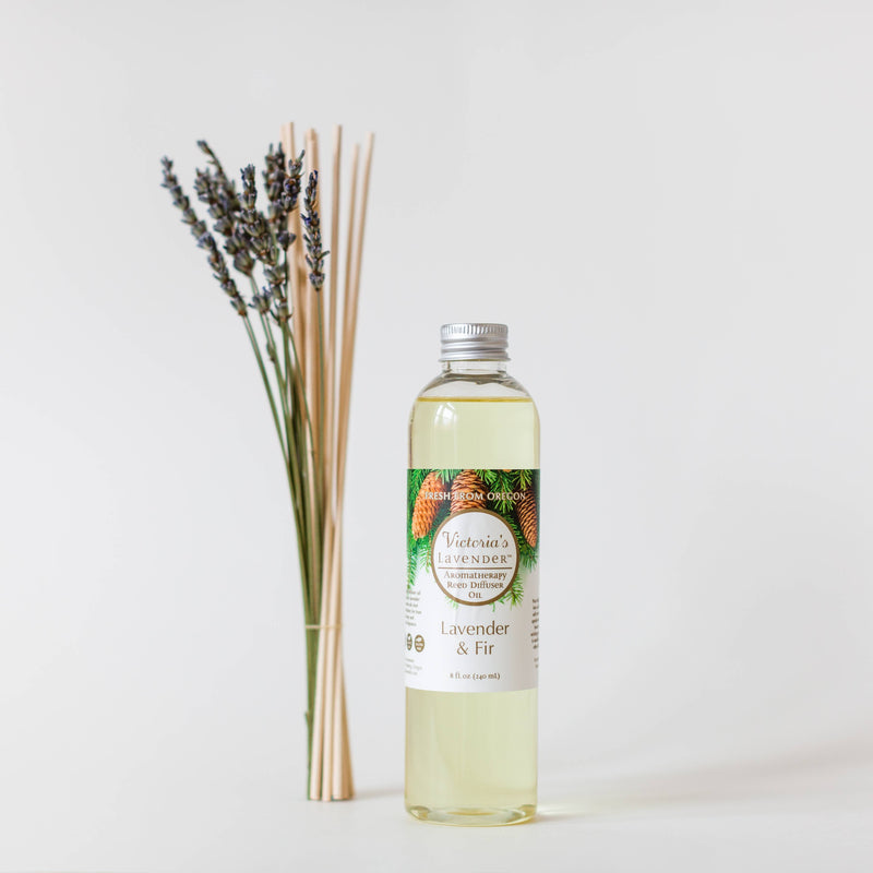A clear bottle of Victoria's Lavender - Lavender & Fir Reed Diffuser Refill Oil 8 oz with a label displaying lavender and fir on a white background, accompanied by lavender sprigs and reed diffuser oil sticks to the side.